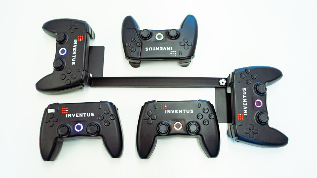 Multiple black Inventus gaming controllers arranged around a mechanical test setup on a white background.
